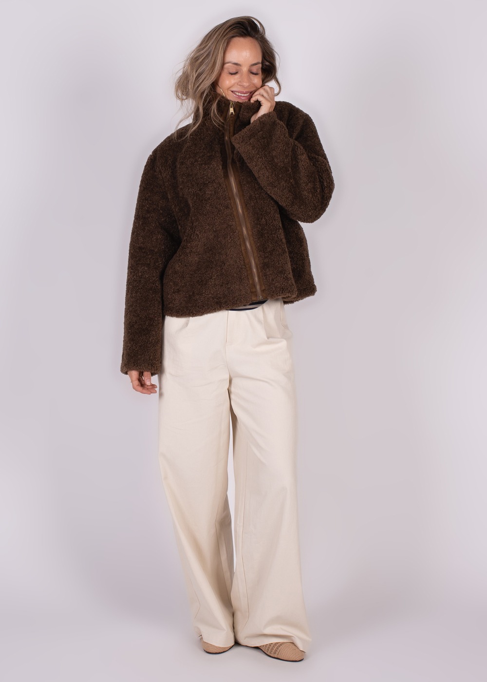 Model in een zachte, bruine Regina jas met rits, gecombineerd met wijde, witte broek, tegen een neutrale achtergrond.