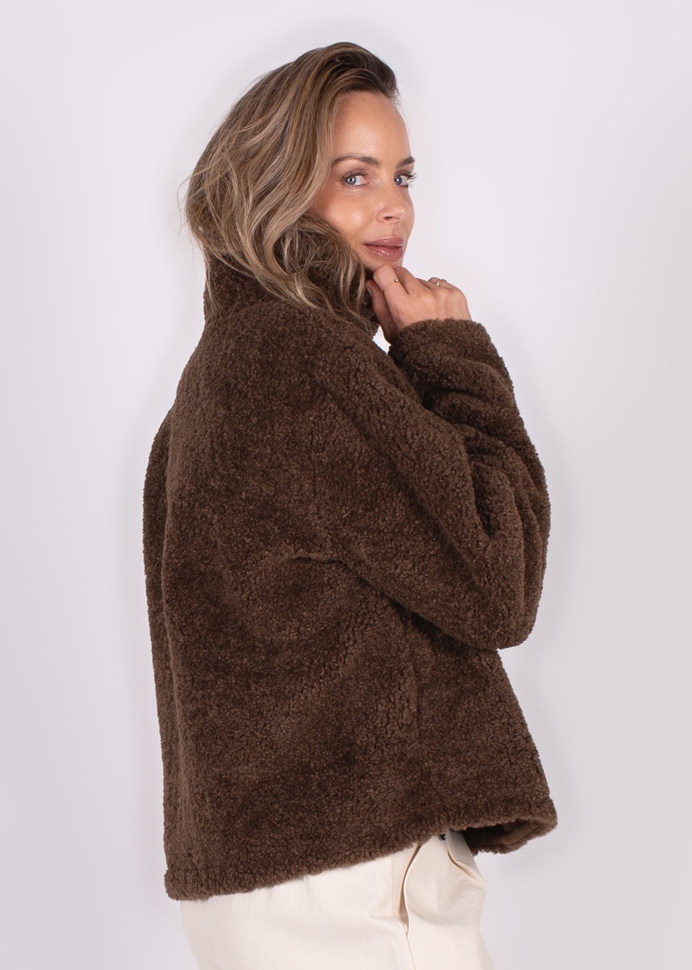 Een vrouw in een bruin, wollig jack met losse kragen poseert zijwaarts, kijkend over haar schouder tegen een neutrale achtergrond.