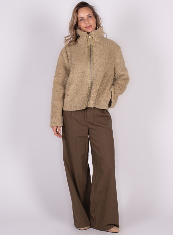 Model draagt een beige Regina jas met opstaande kraag, gecombineerd met wijde bruine broek, voor een witte achtergrond.