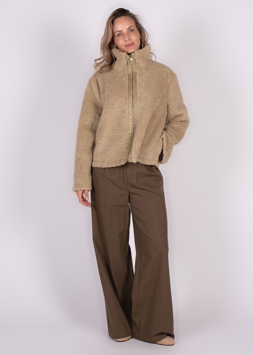 Model draagt een beige Regina jas met opstaande kraag, gecombineerd met wijde bruine broek, voor een witte achtergrond.