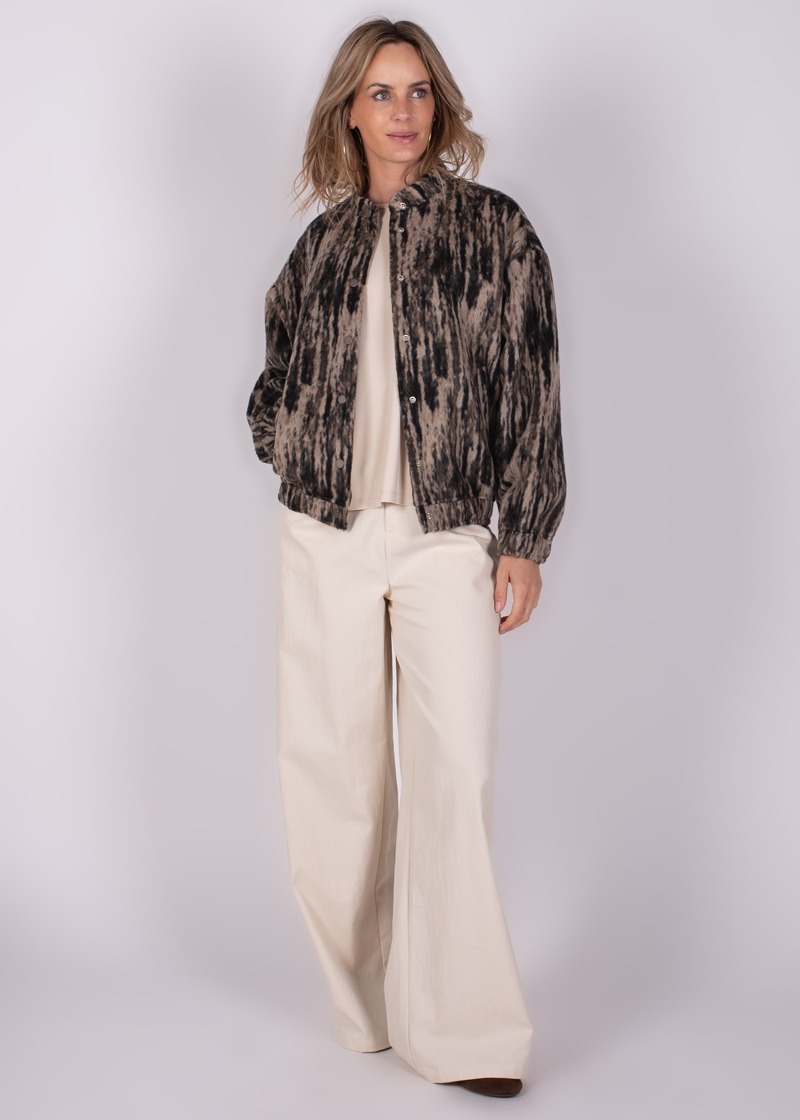Model gekleed in een beige outfit met een kort, bedrukt jasje en wijde broek, gefotografeerd tegen een grijze achtergrond.