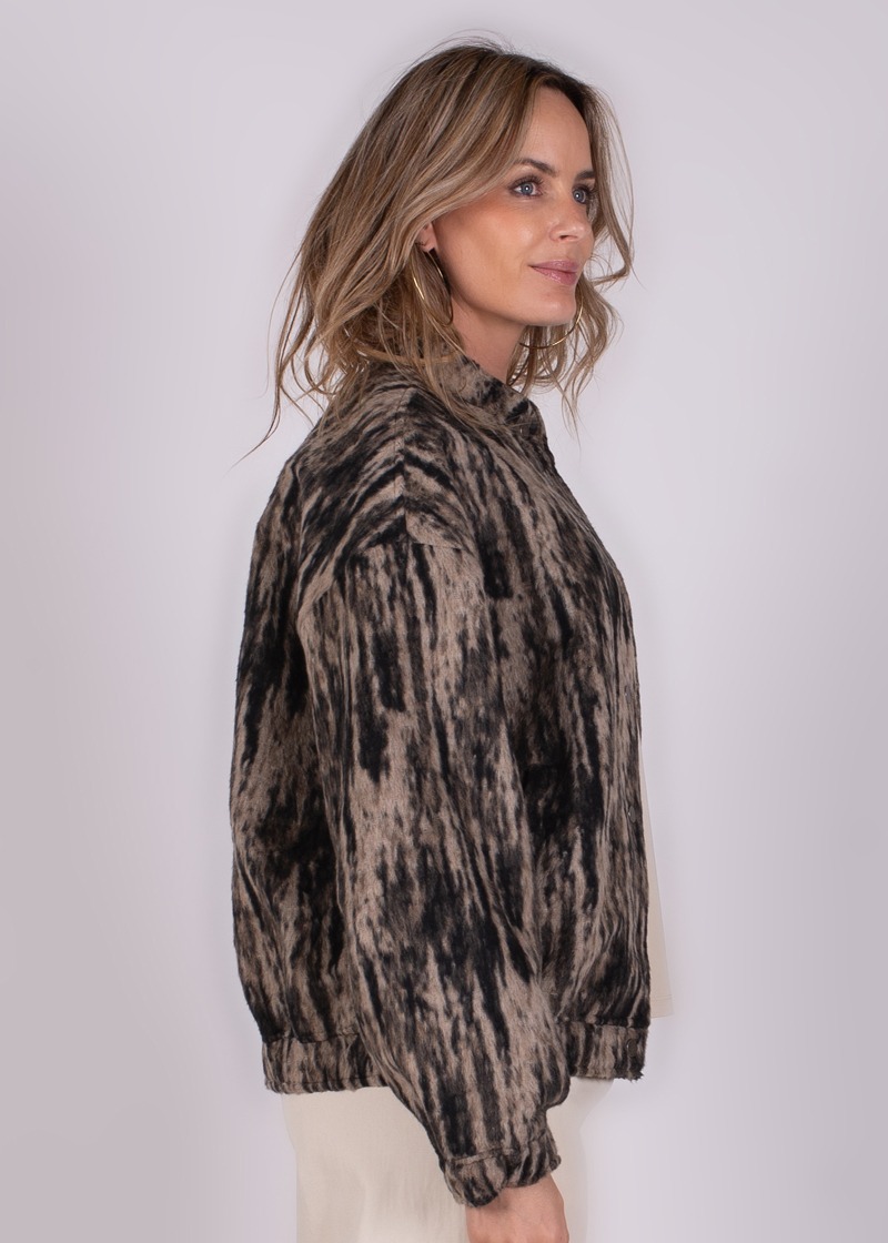 Vrouw in een bruin met zwarte, gemarmerde jas kijkt naar rechts tegen een grijze achtergrond.