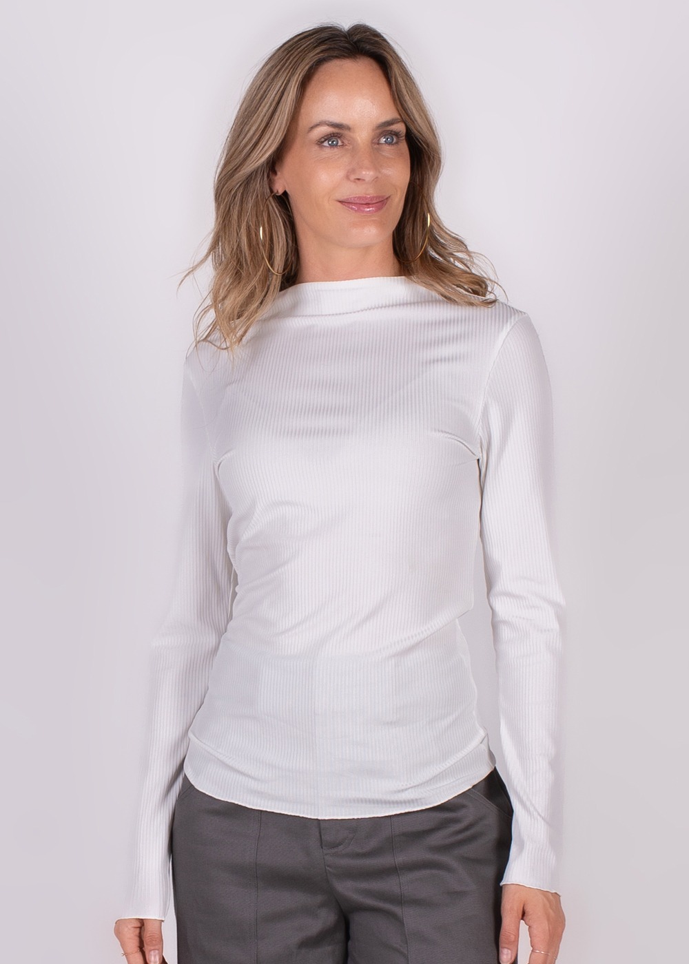 Vrouw met lang haar draagt een witte ribbeltrui en grijze broek, kijkend naar links tegen een neutrale achtergrond.