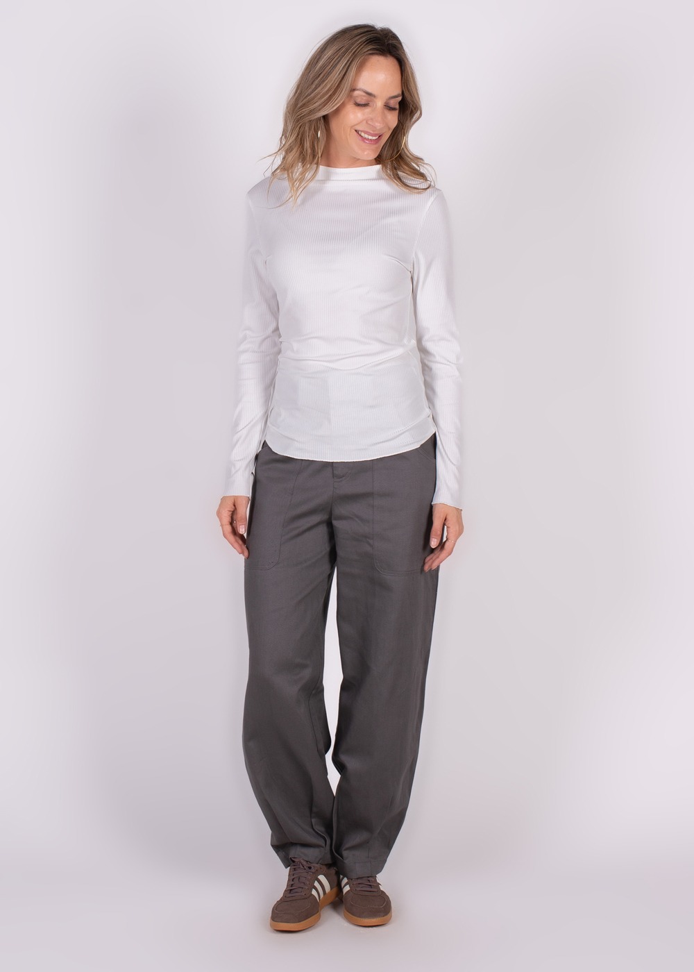 Een lachende vrouw draagt een witte top en grijze broek tegen een neutrale achtergrond.