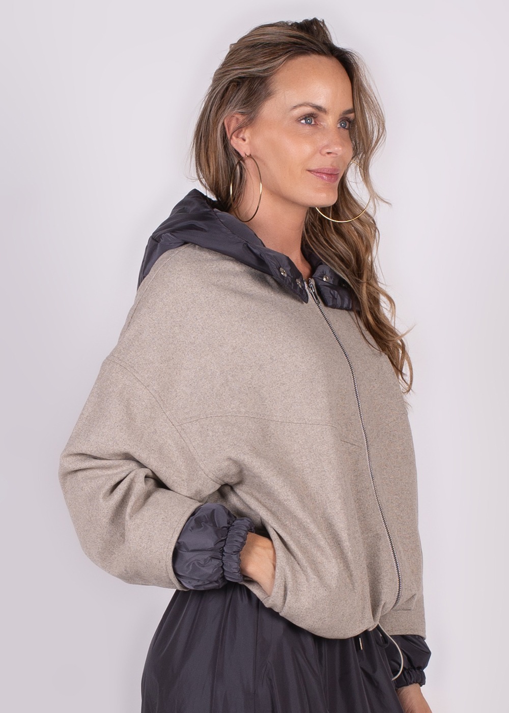 Een vrouw draagt een beige Surat Jacket met een donkere, glanzende capuchon, kijkend naar rechts.