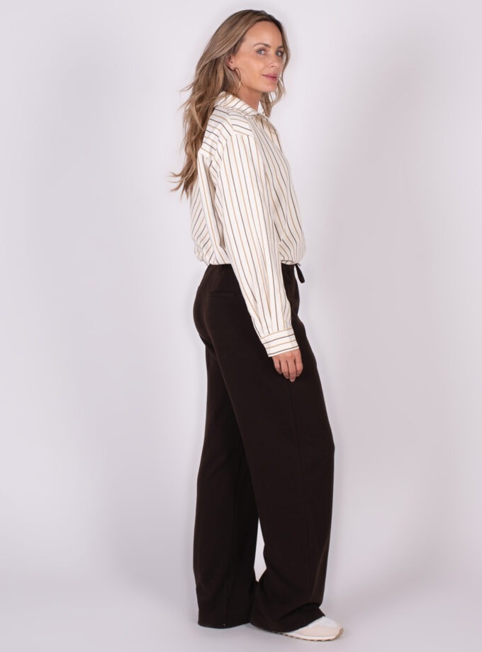 Een vrouw gekleed in een gestreepte blouse en donkere wijde broek staat zijwaarts, met de blik naar links.