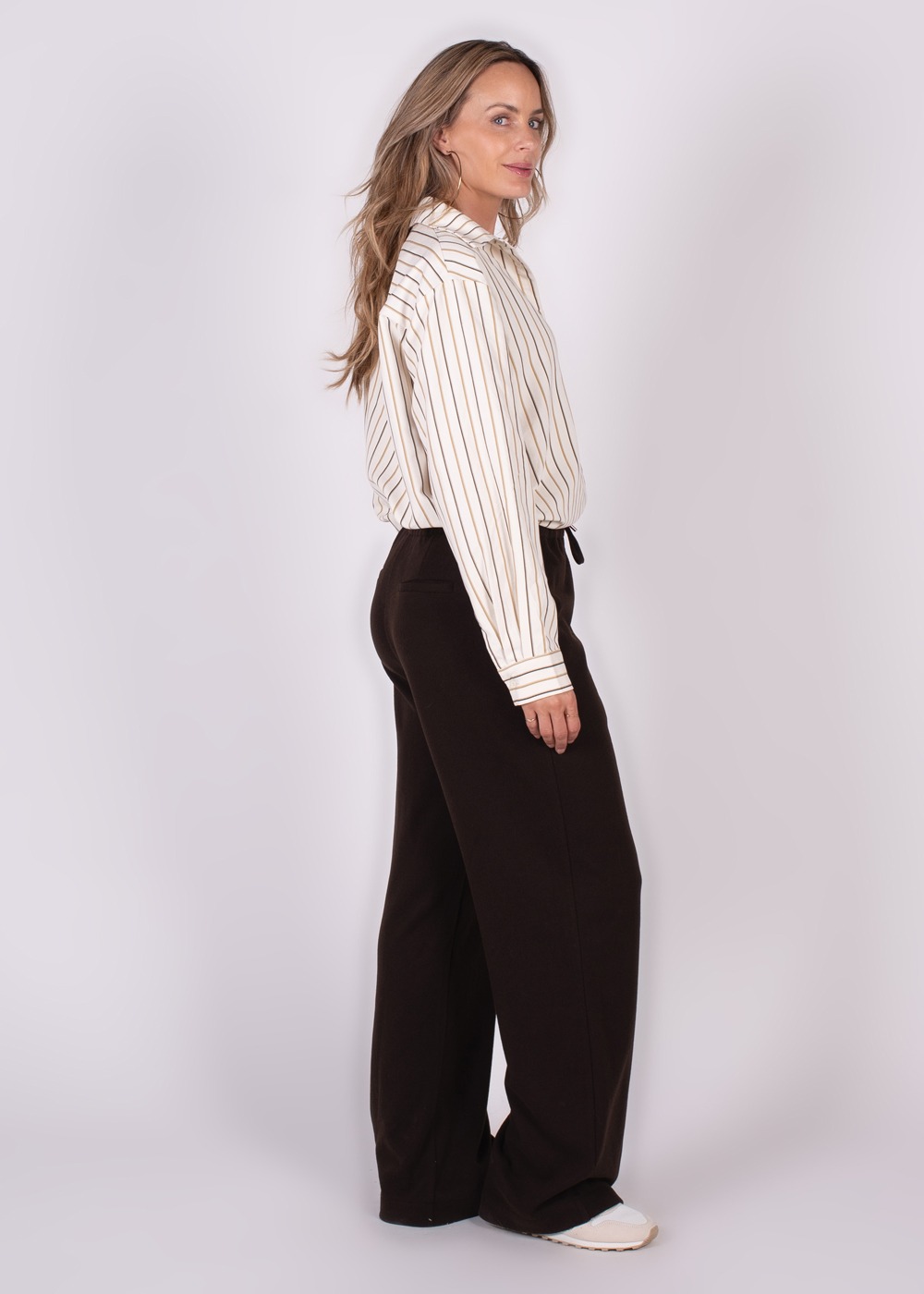 Een vrouw gekleed in een gestreepte blouse en donkere wijde broek staat zijwaarts, met de blik naar links.