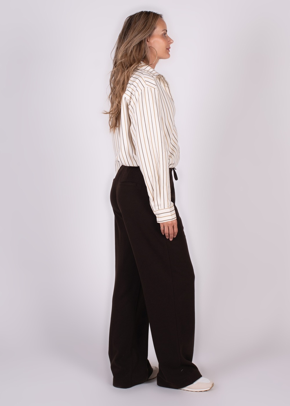 Een model in zijaanzicht draagt een wit gestreepte blouse en zwarte Surat-broek.