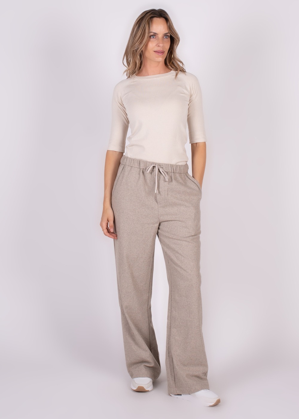 Een vrouw staat in een rechte pose en draagt wijde, beige Surat-broek met een detail op de zak en een lichtbeige shirt.