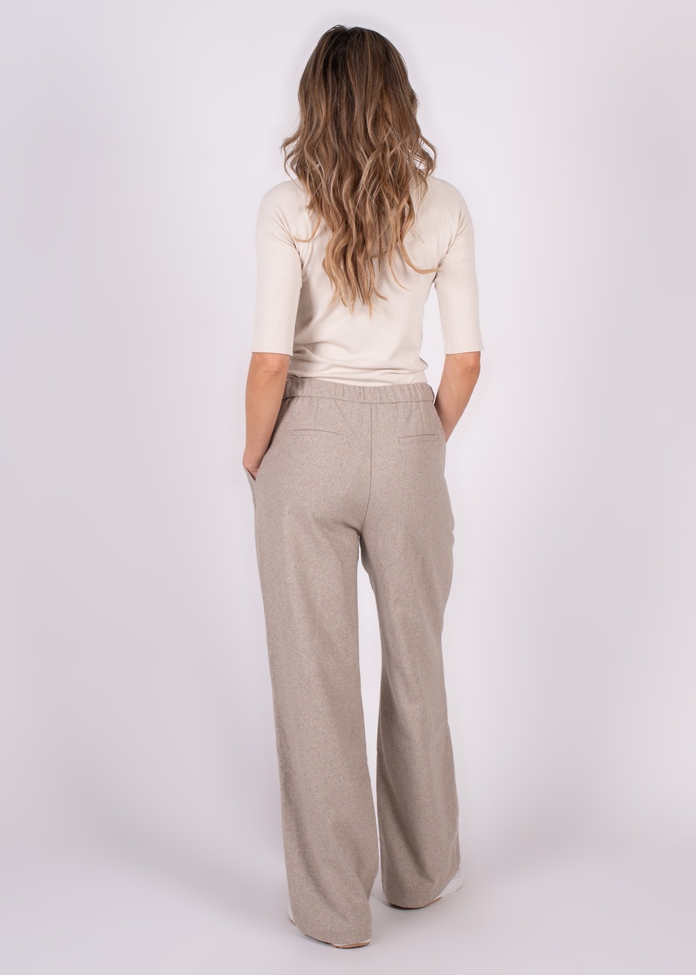 Een persoon met lang golvend haar draagt een beige shirt en grijze wijde Surat-broek met gedetailleerde zakken, gefotografeerd van achteren.