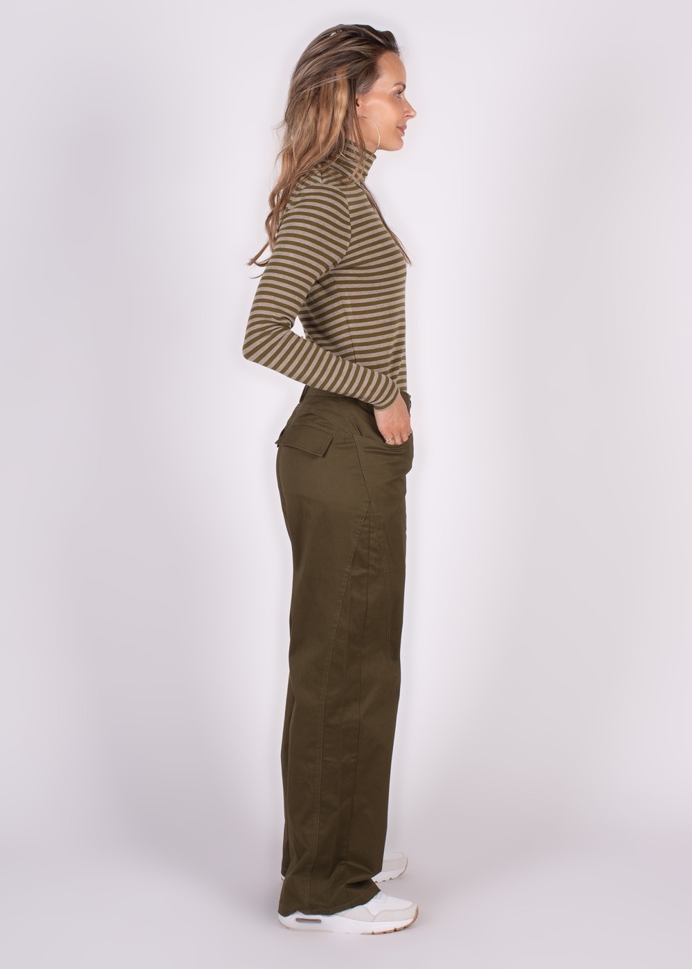 Model poseert zijwaarts in een gestreepte trui en olijfgroene "Vienna pant moss", met witte sneakers.