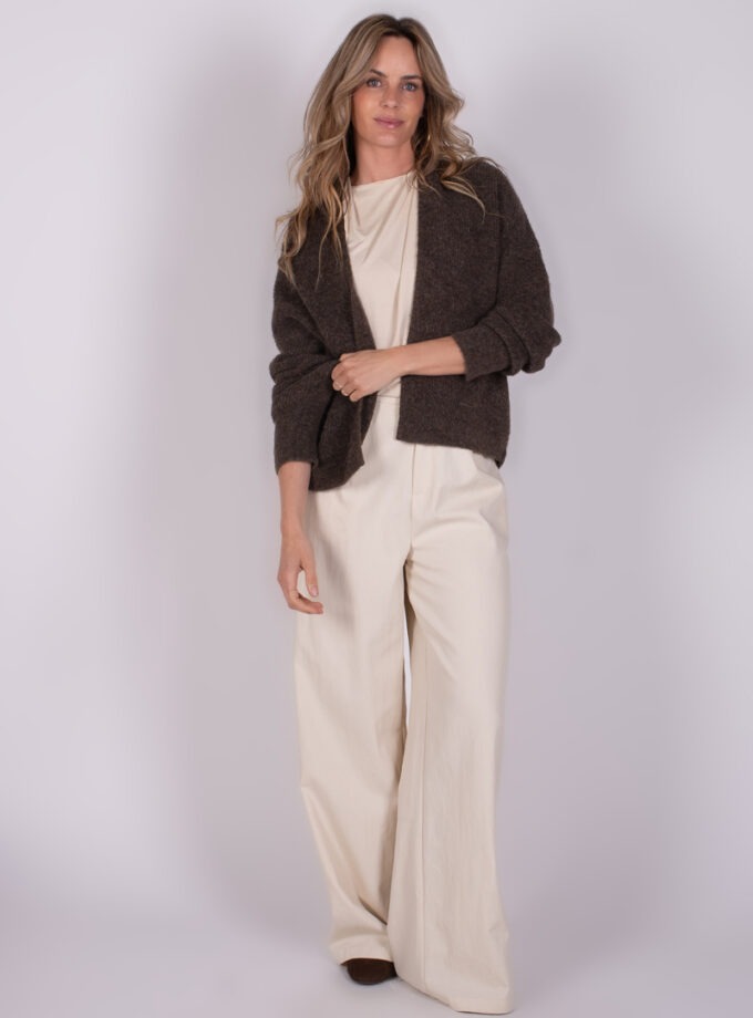 Een vrouw poseert in een bruine "Lamia" cardigan en crèmekleurige wijde broek tegen een neutrale achtergrond.