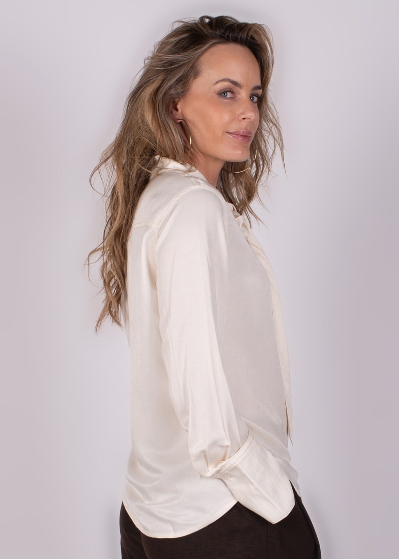 Een vrouw met lang haar en een zijden blouse kijkt over haar schouder in een zijaanzicht tegen een neutrale achtergrond.