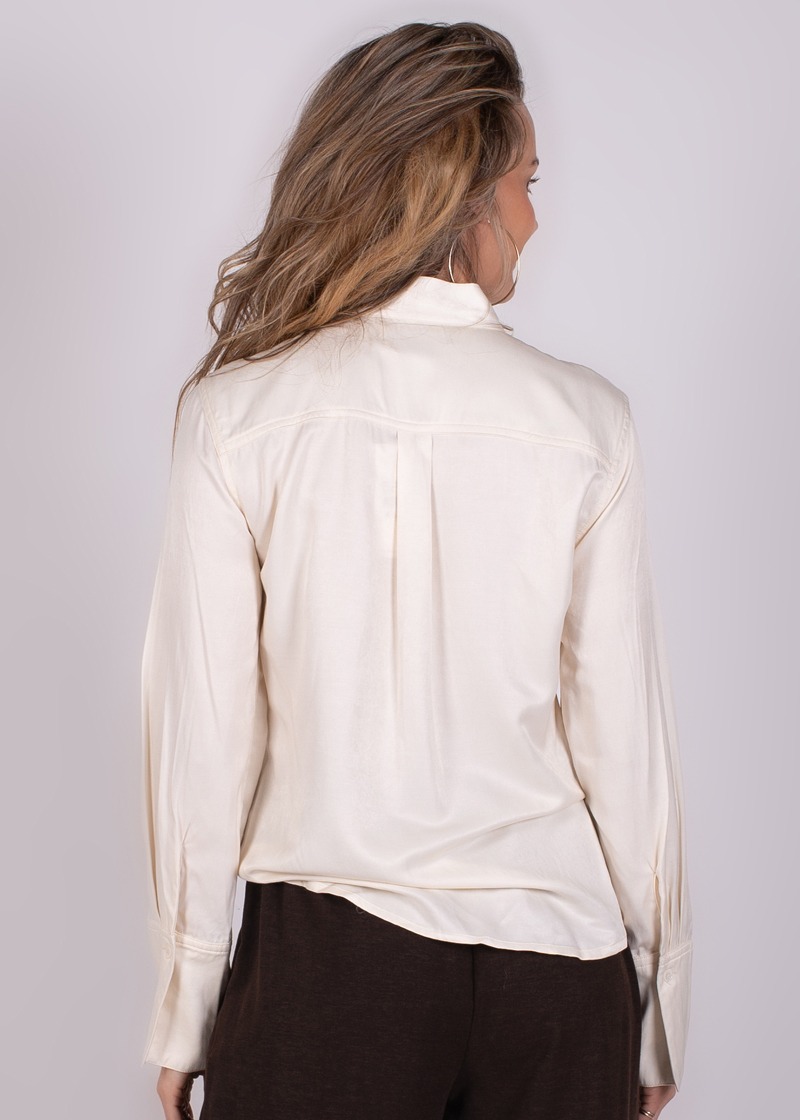 Achteraanzicht van een vrouw in een crèmekleurige blouse en donkere broek tegen een grijze achtergrond.