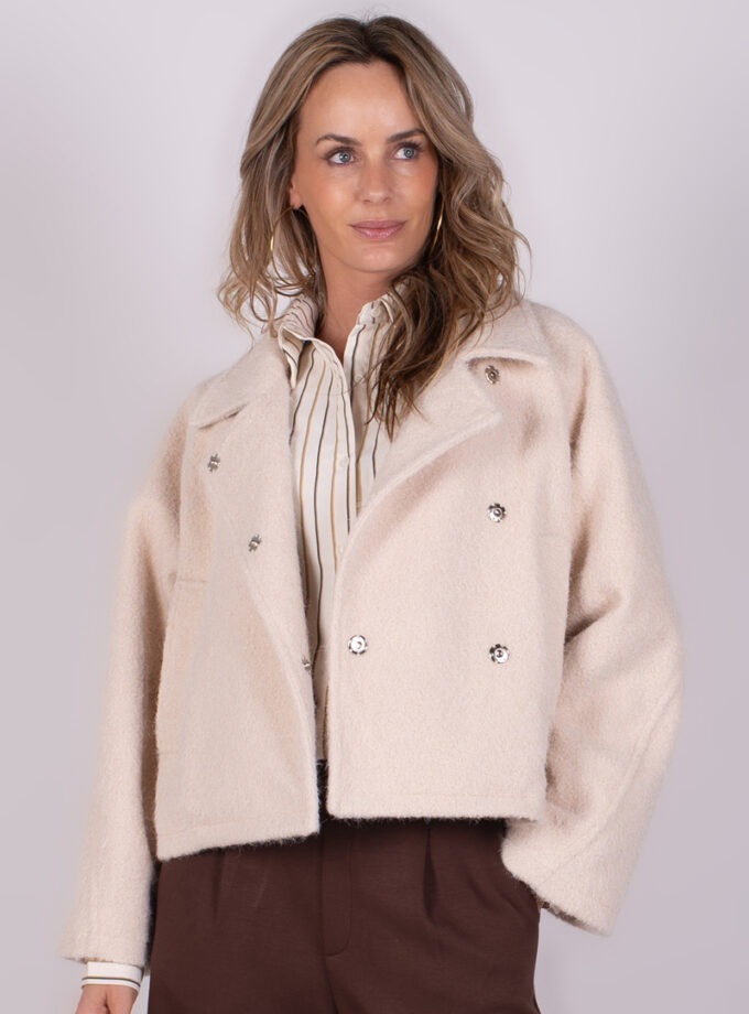 Model draagt een lichtbeige "Matsumoto" jas met dubbele knopen en een gestreepte blouse.